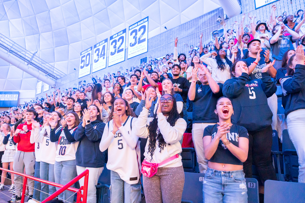 A large group of people are standing and cheering in a stadium. Many are wearing UConn jerseys and shirts, with the number 5 being prominent. The background shows retired jersey numbers 50, 34, and 32 hanging on the wall. The crowd appears to be very enthusiastic and engaged in the event.