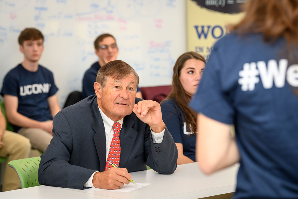 Classroom scene with a person in a suit taking notes, others in UCONN shirts seated behind, and a whiteboard with diagrams in the background.