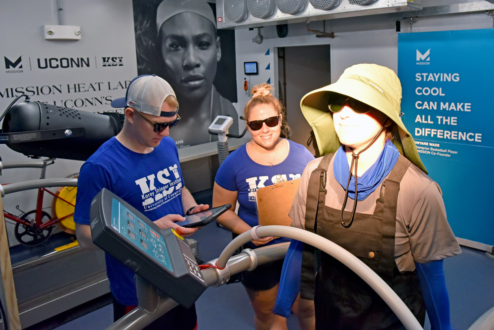 Three people in a lab, one on a treadmill wearing a large hat, while two others observe and take notes. Lab equipment and a sign reading 'STAYING COOL CAN MAKE ALL THE DIFFERENCE' are in the background.