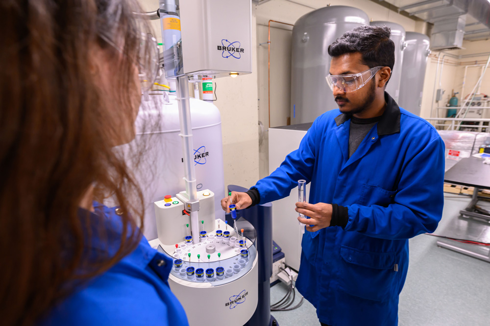 Two individuals in blue lab coats are working with a Bruker scientific instrument in a laboratory setting. One person is holding a test tube while the other observes. The background includes various laboratory equipment and storage tanks.